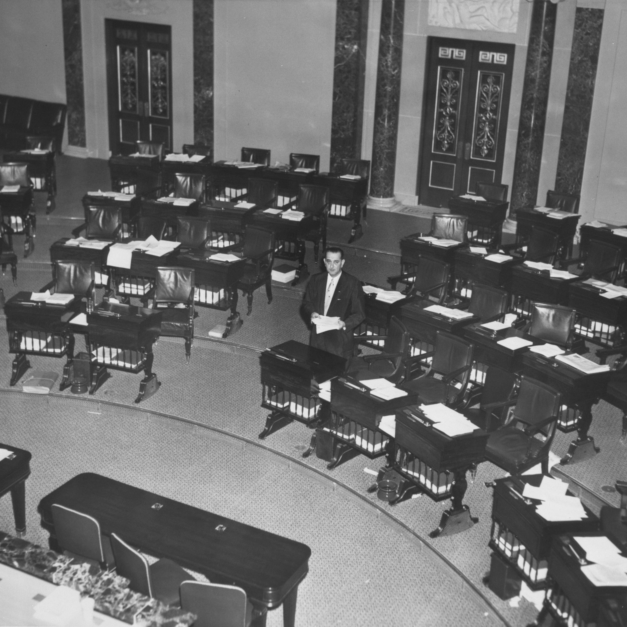A wide shot from above. Senator Lyndon B. Johnson, alone in the senate chamber, cleans out his desk. 